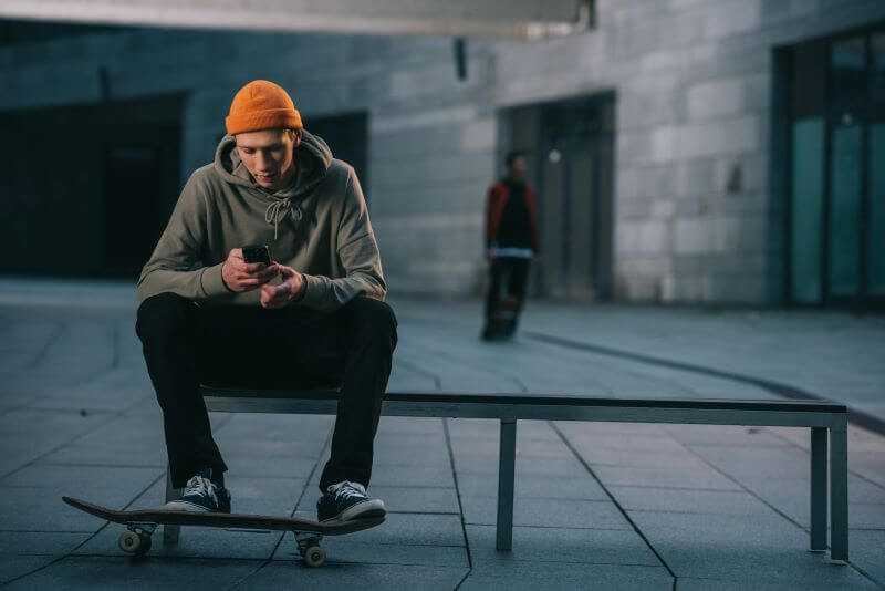skateboarder in modern streetwear sitting on bench and using smartphone